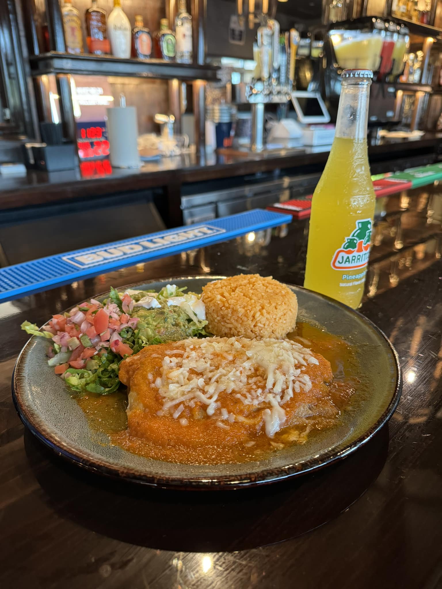 A plate of chicken enchiladas with a side of rice, salsa, and guacamole. The dish is topped with cheese and a red sauce. A bottle of Jarritos pineapple soda can be seen in the background.