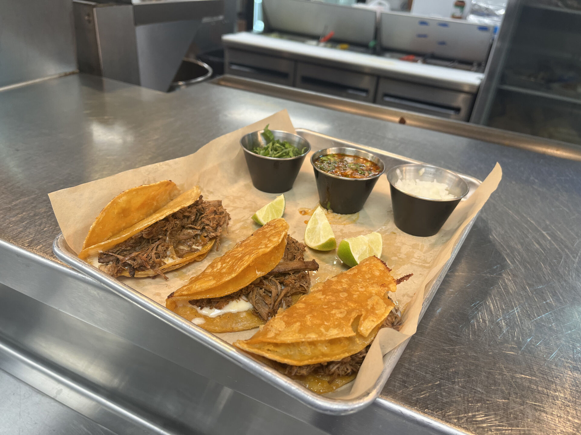 Three beef tacos on a tray with cilantro, salsa, and lime wedges.